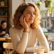 Woman sitting at a cafe table with a cup of coffee, looking out the window. Gold vermeil wire wrap bracelet can be made in a variety of birthstones.