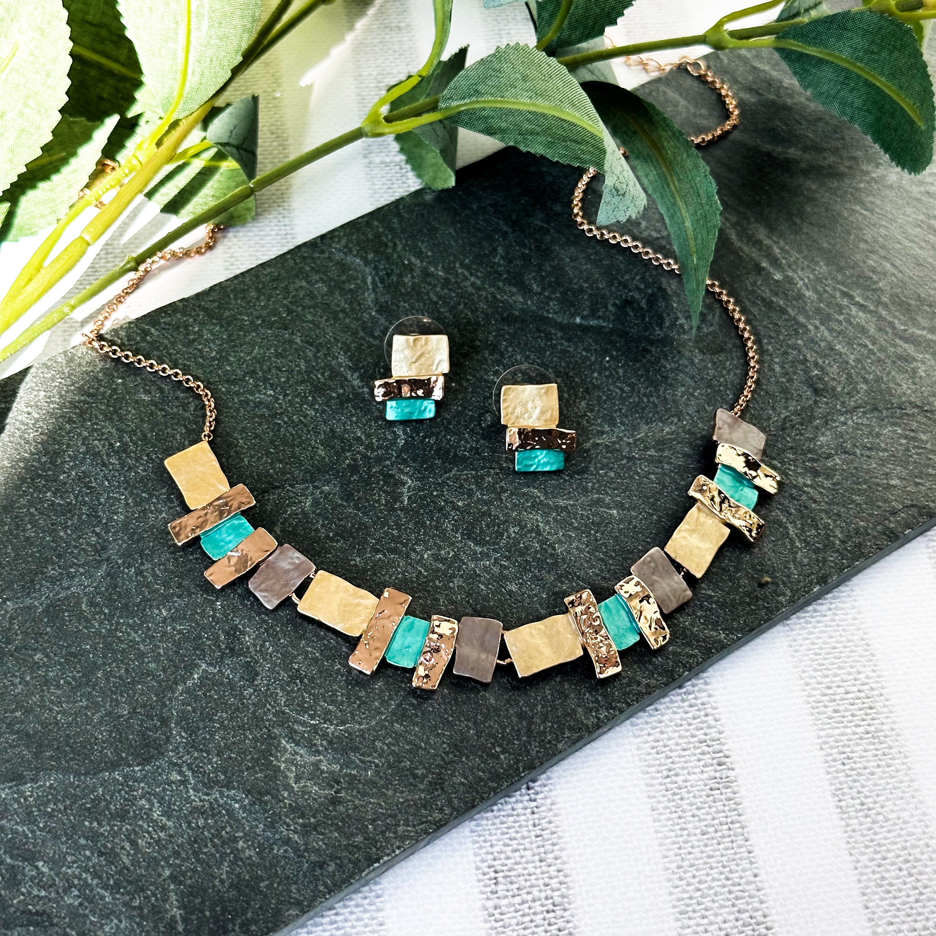 Adjustable yellow gold necklace with a cable chain and pendant made of textured metal rectangle and squares painted in yellow peach and brown aqua enamel sits on a white plate on a peach stone background set of necklace and earrings