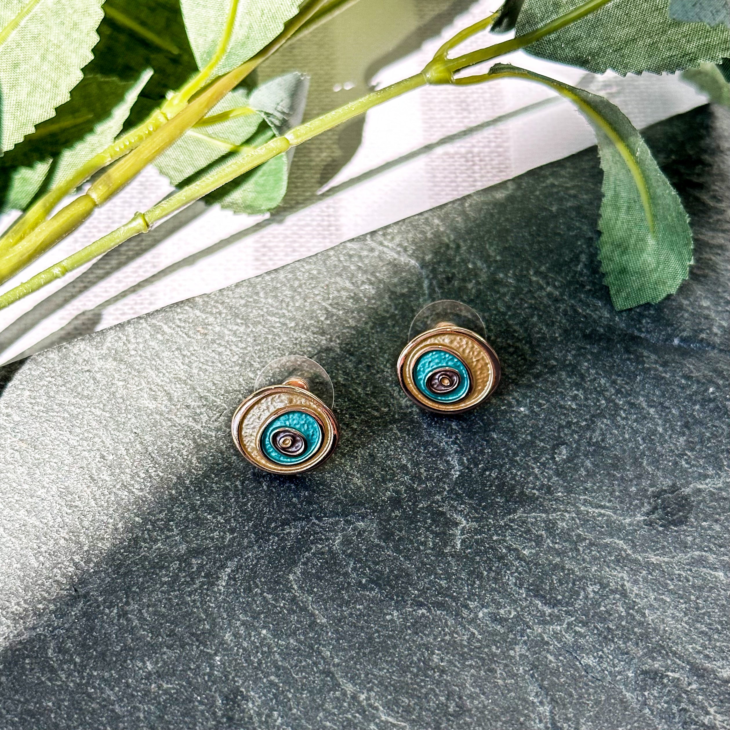 A pair of gold, teal and purple swirl stud earrings rest on a black granite background with leafy green stalks behind them. Boho summer style earrings and necklace set, gold and green earrings, abstract evil eye jewelry, subtle evil eye jewelry