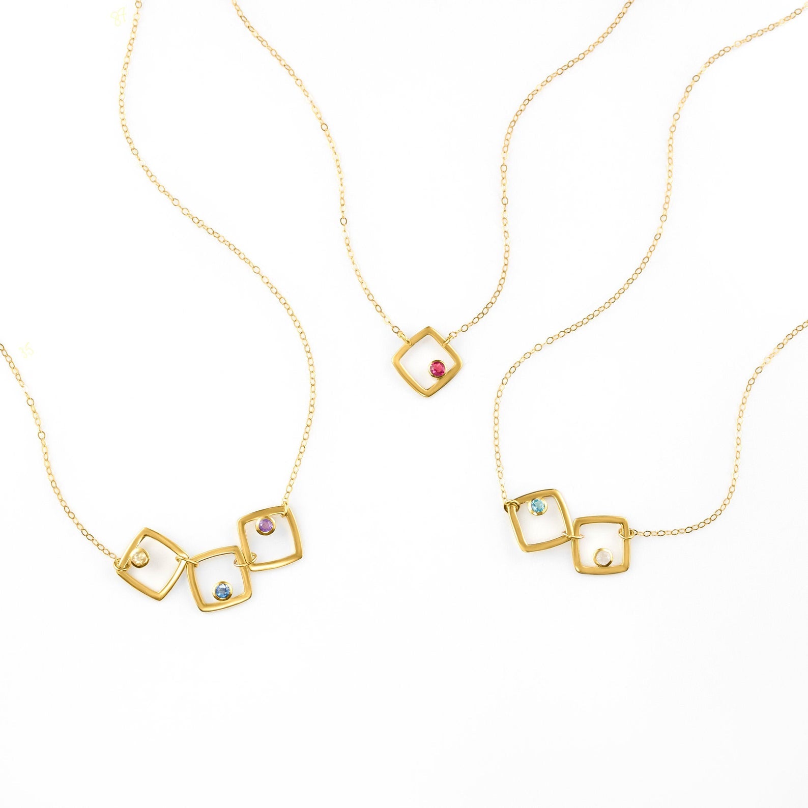 Trio of gold filled square geometric necklaces. First necklace has citrine, kyanite and purple amethyst birthstones. Center necklace features a single ruby birthstone. Final necklace has blue topaz and moonstone birthstones. 