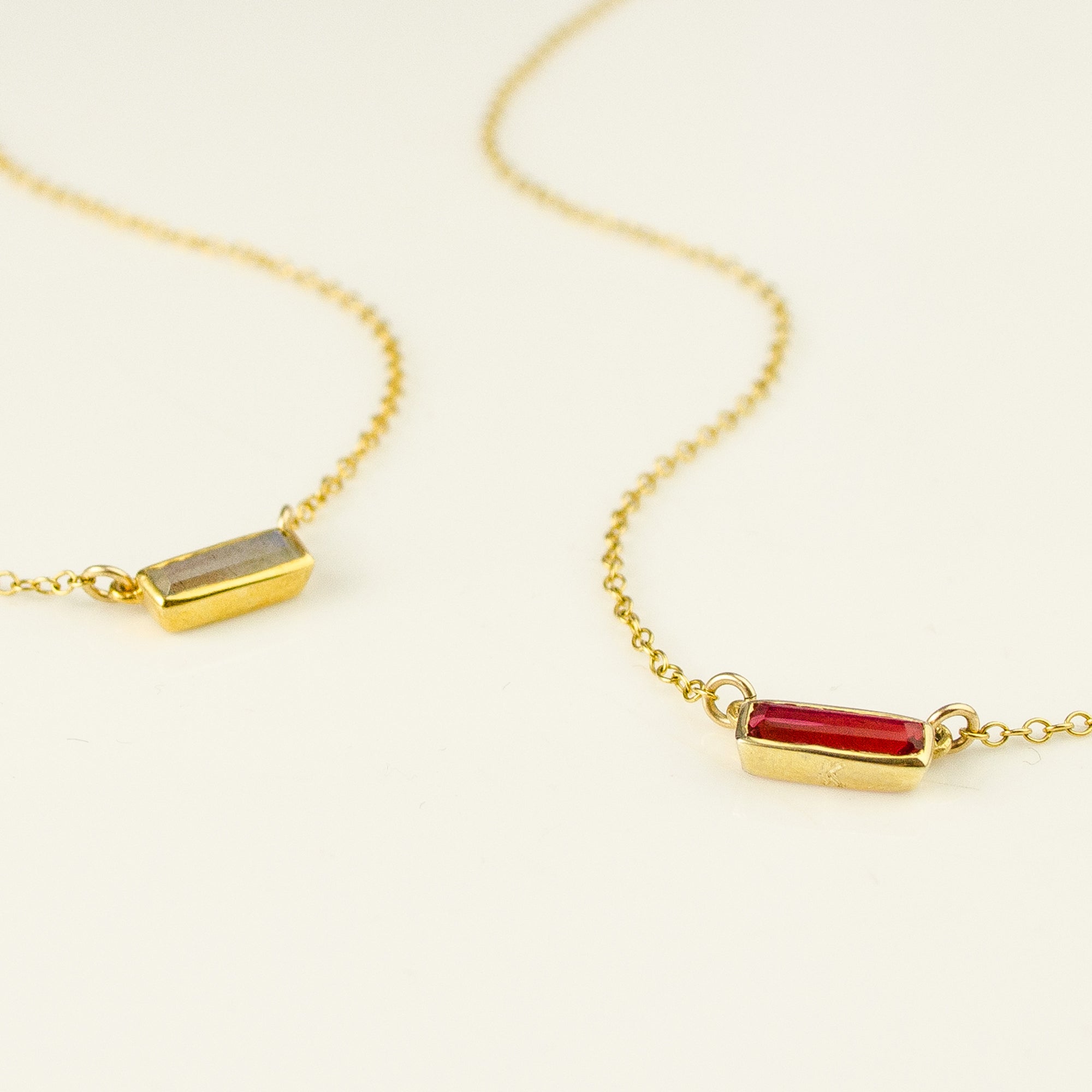 Two gold filled tiny adira bar necklaces on white background featuring labradorite and garnet birthstones. Garnet setting has an engraved heart emoji. 