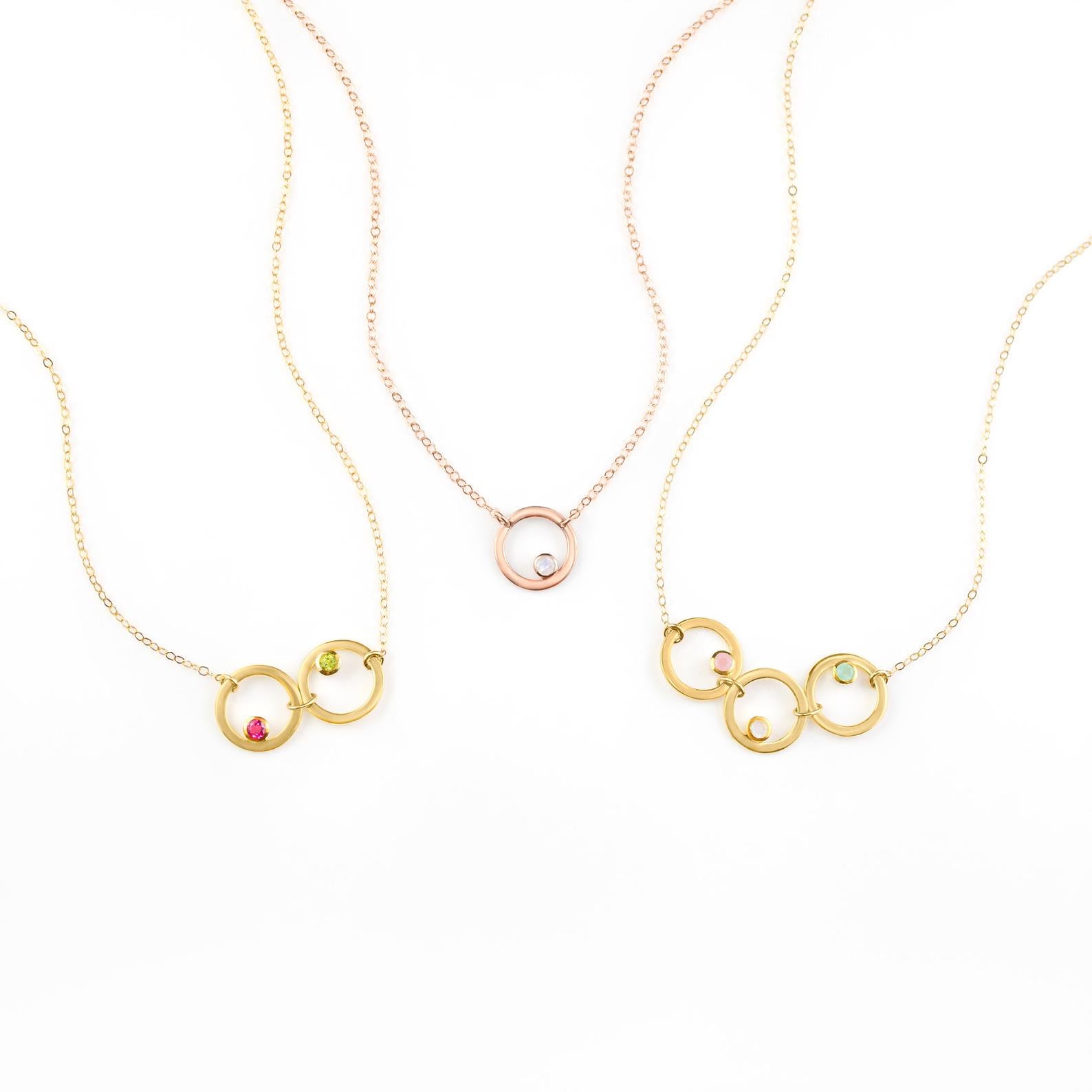 Collection of open circle eden necklaces from left to right. One gold filled necklace with ruby and peridot birthstones, one rose gold necklace with moonstone, and one gold necklace with pink chalcedony, moonstone and aqua chalcedony birthstones. 