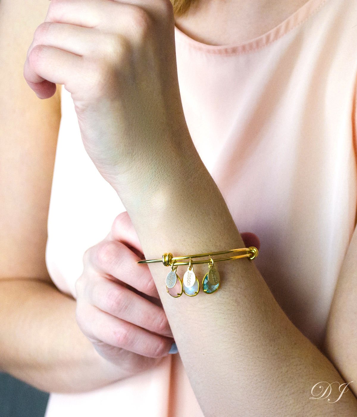Model is wearing a gold bangle with three name tags and three birthstones including pink chalcedony, moonstone and blue topaz