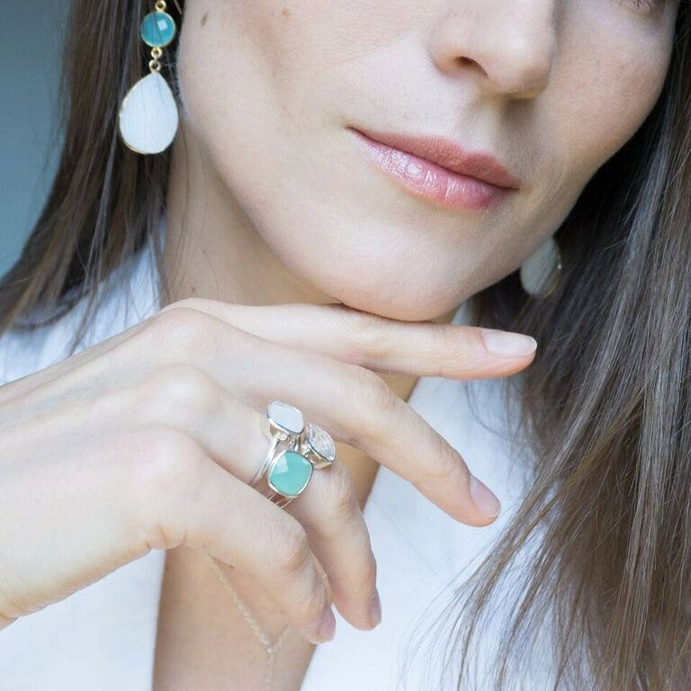 Model is stylishly stacking three square cushion bezel rings in moonstone, clear quartz and aqua chalcedony in stamped 925 sterling silver. 