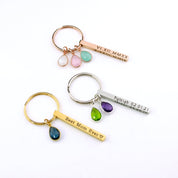 Flat lay of several family keychains in gold, silver, and rose gold, each with engraved 3D bars and different birthstone charm combinations to represent kids or grandkids. The photo illustrates customization options—single or multiple stones, various fonts, and four-side engraving—making a versatile name bar keychain for Mother’s Day, baby showers, and everyday gifts for mom.