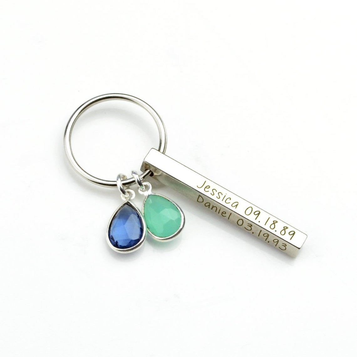 Minimal product view of a silver mom keychain photographed on a bright white background. A dainty 3D bar is engraved with a name, paired with two bezel-set birthstones in ocean hues. Lightweight and durable for daily use, this personalized keychain can include additional stones for siblings and custom text on up to four sides for a heartfelt, practical gift.