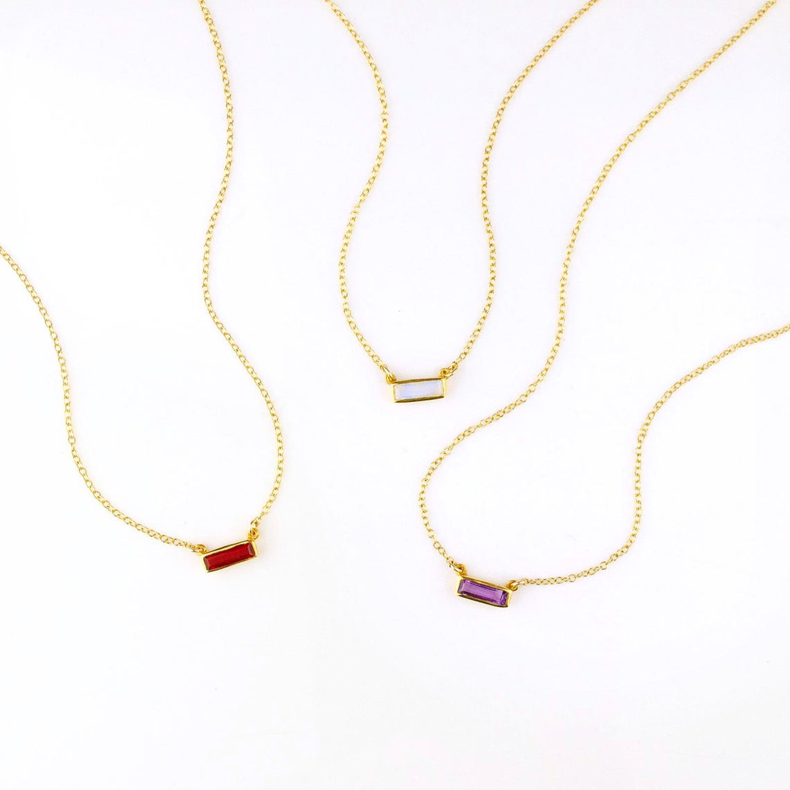 Three tiny gold filled adira bar necklaces on white background featuring garnet, moonstone and purple amethyst birthstones. 
