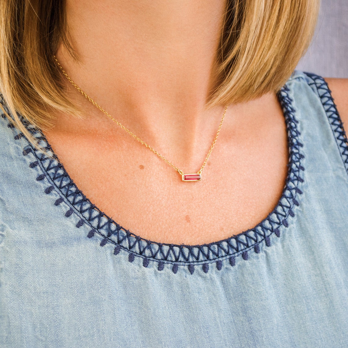 Model in blue top is wearing a single gold filled tiny ruby birthstone adira bar necklace. 