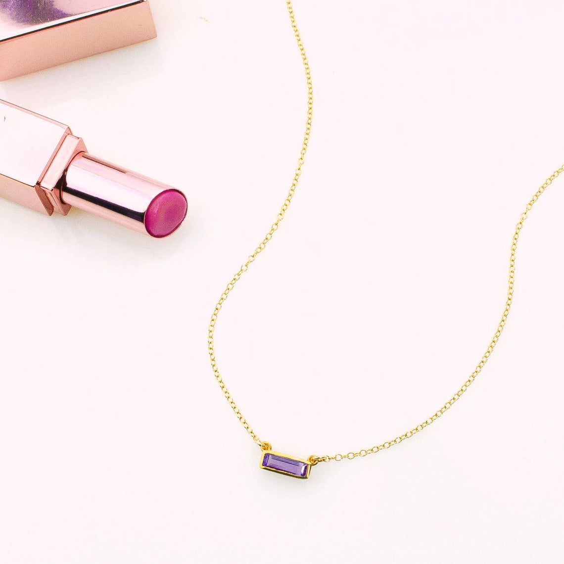 A gold filled tiny adira bar necklace featuring a purple amethyst birthstone on white background with lipstick - great mother's day, birthday, celebration or anniversary gift.